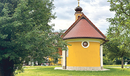 Eine Kapelle (Gelb) in einem Garten umgeben von Bäumen.