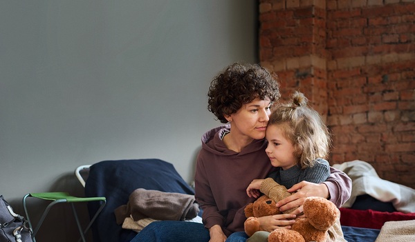 Young woman giving hug to her cute little son with brown soft teddybear while both sitting on sleeping place prepared for refugees