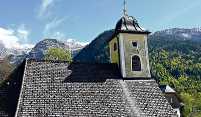 Die katholische Pfarrkirche vor der malerischen Bergkulisse. Beim Kirchturm sind die renovierungsbedürftigen Schäden sichtbar.  