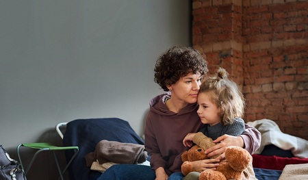 Young woman giving hug to her cute little son with brown soft teddybear while both sitting on sleeping place prepared for refugees