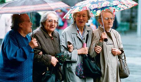 Vier Seniorinnen spazieren mit Regenschirmen bei Regenwetter Arm in Arm auf der Stra?e